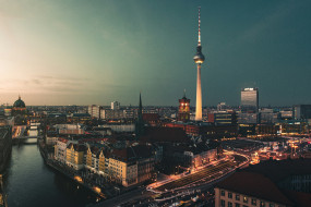 Stadtansicht von Berlin bei Dämmerung mit Fluss, Kuppel, Fernsehturm und beleuchteten Straßen. Keine Personen sichtbar.