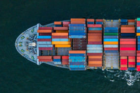 Containerschiff aus der Vogelperspektive, beladen mit bunten Containern, im dunklen Wasser mit Wellen am Heck.