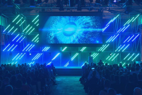 Veranstaltung in großem Raum mit sitzenden Personen, großer Leinwand mit blauem Lichtkreis und geometrischen Lichtinstallationen.