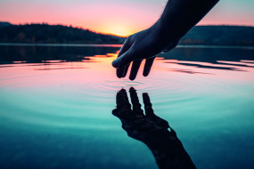 Hand über Wasser, Finger berühren die Oberfläche und erzeugen Wellen. Landschaft mit Bäumen und sanfter Himmel im Hintergrund.