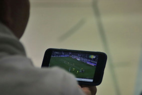 Mann mit hellem Pullover schaut auf Smartphone-Bildschirm mit Fußballszene und Punktestand 0:0.