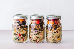 Drei Schraubgläser mit Quinoa, buntem Gemüse und kleinen Tomaten auf einer flachen Oberfläche. Metallene Schraubverschlüsse.