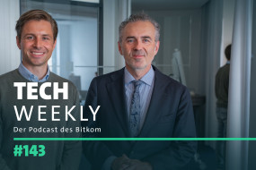 Zwei Personen stehen nebeneinander. Links: hellbraune Haare, grauer Pullover, blaues Hemd. Rechts: graue Haare, dunkler Anzug, hellblaue Krawatte. Hintergrund: helle Wände, Fenster. Text: "TECH WEEKLY", "