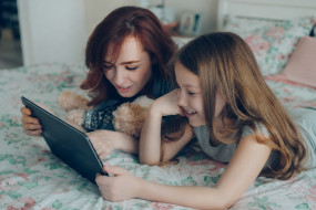 Mutter und Kind liegen auf einem Bett, schauen auf ein Tablet. Kind hat einen Teddybären neben sich.