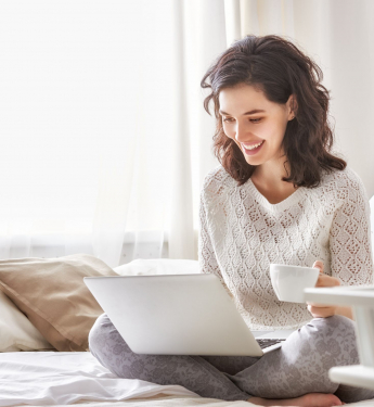 Frau sitzt mit Laptop und einer Tasse Kaffee im Bett