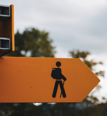 Gelbes Schild mit Wanderwegsymbol und Pfeil nach rechts, befestigt an grauem Metallpfosten. Unscharfer Hintergrund mit Bäumen.