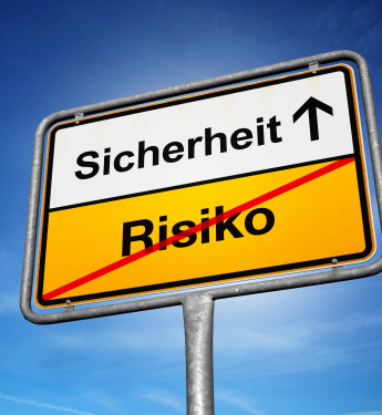 Ein gelb-weißes Straßenschild mit schwarzer Schrift steht vor einem strahlend blauen Himmel. Das obere, weiße Feld zeigt das Wort „Sicherheit“ mit einem nach oben zeigenden Pfeil. Das untere, gelbe Feld enthält das Wort „Risiko“, das mit einem roten diagonalen Strich durchgestrichen ist. Das Schild erinnert an Ortsschilder und symbolisiert die Bewegung von Risiko hin zu mehr Sicherheit.