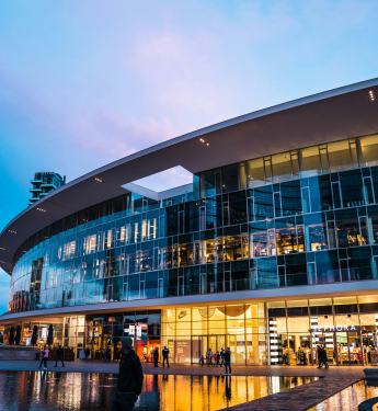 Ein großes, modernes Einkaufszentrum mit einer geschwungenen Glasfassade reflektiert das warme Licht der Geschäfte im Inneren. Die untere Etage beherbergt bekannte Marken wie Nike, Sephora und Under Armour, deren beleuchtete Logos sichtbar sind. Vor dem Gebäude befindet sich ein flacher Wasserbereich mit kleinen Lichtern. Mehrere Personen spazieren entlang der Promenade, einige tragen Kapuzenpullover oder Jacken. Der Himmel zeigt eine Mischung aus Blau und Rosa, da die Sonne untergeht.