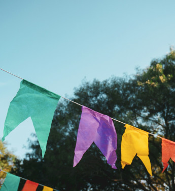Bunte Wimpelgirlanden hängen im Freien vor blauem Himmel und grünen Bäumen.