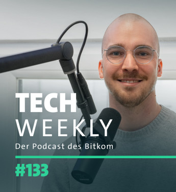 Mann mit Glatze und Brille lächelt vor Mikrofon, trägt grauen Pullover. Hintergrund: helle Wände und Fenster.