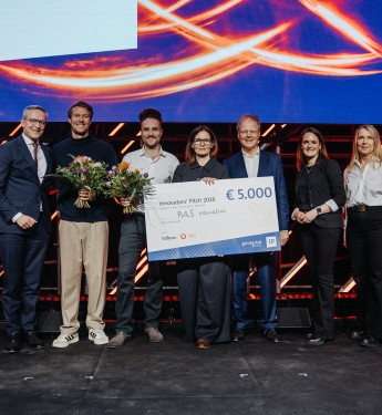 Gruppe von fünf Personen auf einer Bühne, Frau mit Scheck über € 5.000, leuchtende Muster im Hintergrund.