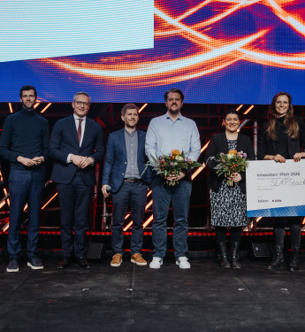 Sechs Personen in formeller Kleidung auf einer Bühne, eine hält einen Scheck über € 5.000, andere mit Blumensträußen.