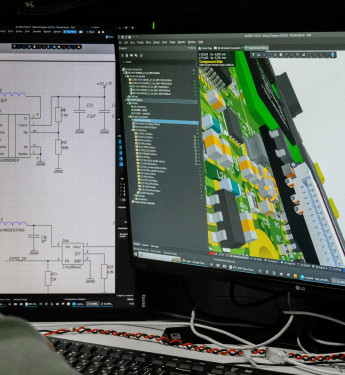 Person von hinten am Computer, links ein Schaltbild, rechts eine 3D-Leiterplatte, Kabel und Werkzeuge auf dem Tisch.