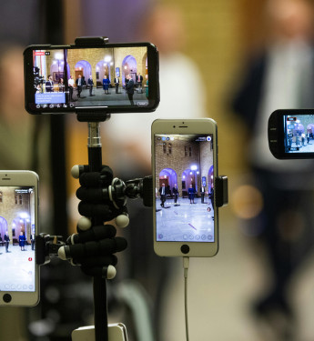 Mehrere Smartphones und eine Kamera sind auf Stativen ausgerichtet und filmen eine Veranstaltung in einem Innenraum. Auf den Displays ist eine Gruppe von Personen zu sehen, die in einem beleuchteten Raum mit Bögen im Hintergrund stehen. Die Geräte übertragen oder zeichnen die Szene parallel aus verschiedenen Perspektiven auf, während der Hintergrund unscharf bleibt.