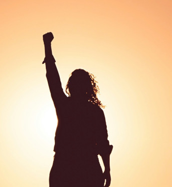 Silhouette einer Frau mit erhobener Faust vor einem orangefarbenen Sonnenuntergangshintergrund.