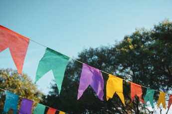 Bunte Wimpelgirlanden hängen im Freien vor blauem Himmel und grünen Bäumen.