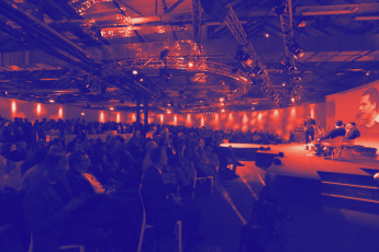 Große Veranstaltungshalle mit Publikum, Bühne und Podium, zwei Personen sitzen, warmes Licht, projiziertes Bild im Hintergrund.