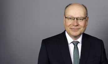 Frank Wetzel, ein Mann mit Glatze und Brille, lächelt in einem dunklen Anzug vor einem grauen Hintergrund.