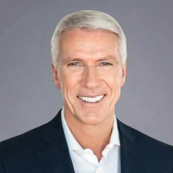 Ralf Wintergerst, Mann mit grauen Haaren, dunklem Anzug und weißem Hemd, lächelt freundlich vor grauem Hintergrund.