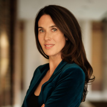 Frau in dunkelblauem Anzug mit verschränkten Armen, lächelt und blickt in die Kamera, unscharfer heller Innenraum im Hintergrund.