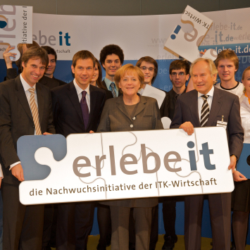 Scheer hält ein Schild mit der Aufschrift "Erlebe IT", u.a. gemeinsam mit Angela Merkel.