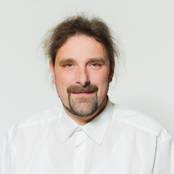 Mann mit langen, lockigen Haaren und Bart, trägt weißes Hemd, lächelt in die Kamera, hellfarbiger Hintergrund.