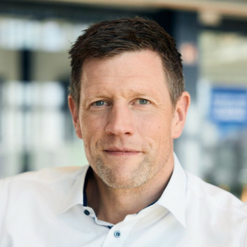 Mann mit kurzen, dunklen Haaren und Bart in weißem Hemd, schaut neutral in die Kamera, unscharfer Hintergrund.