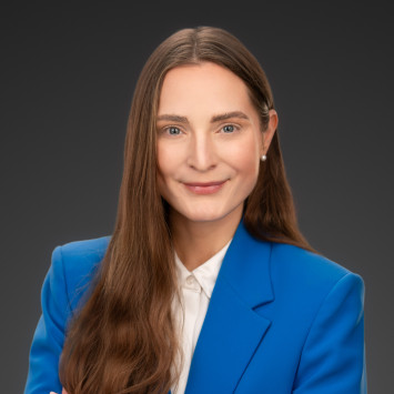 Person mit langen, braunen Haaren in blauem Blazer und weißem Hemd, vor dunklem Hintergrund, lächelt leicht.