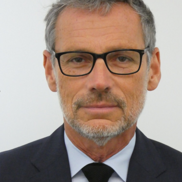 Mann mit grauen Haaren und Bart, schwarzer Brille, dunklem Anzug, hellblauem Hemd, schwarzer Krawatte, schaut in die Kamera.