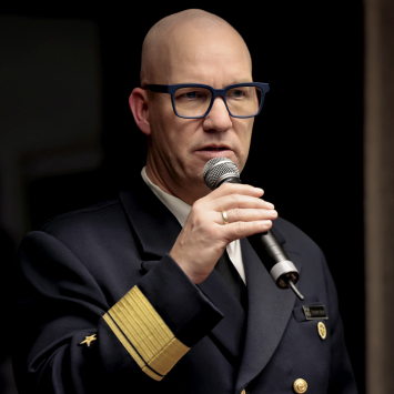 Mann in Uniform mit goldenen Schulterstücken spricht ins Mikrofon, trägt Brille mit blauem Gestell. Dunkler, unscharfer Hintergrund.