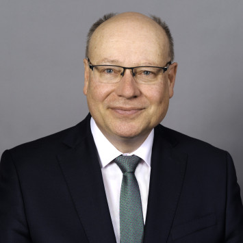 Frank Wetzel lächelt in die Kamera, trägt einen dunklen Anzug und eine gemusterte Krawatte. Grauer Hintergrund.