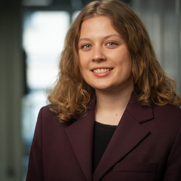 Junge Frau mit lockigem, schulterlangem Haar in dunkelviolettem Blazer, lächelt vor unscharfem, hellem Hintergrund.