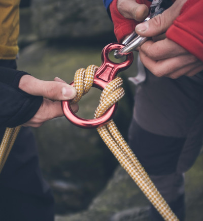 Zwei Hände halten einen roten Karabinerhaken und ein gelbes Seil. Unscharfe graue Felsen im Hintergrund.
