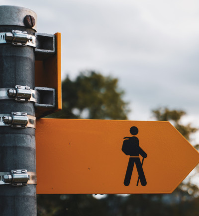 Gelbes Schild mit Wanderwegsymbol und Pfeil nach rechts, befestigt an grauem Metallpfosten. Unscharfer Hintergrund mit Bäumen.