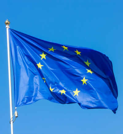 Eine blaue Flagge mit zwölf goldenen Sternen in einem Kreis weht an einem weißen Fahnenmast vor einem klaren blauen Himmel. Die Flagge der Europäischen Union ist leicht gewellt, und die goldenen Sterne heben sich deutlich vom blauen Hintergrund ab. Der Mast hat eine goldene Spitze, und am unteren Teil ist eine Seilvorrichtung zur Befestigung der Flagge zu sehen.