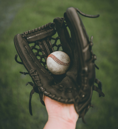 Eine Hand hält einen schwarzen Baseballhandschuh mit sichtbaren Nähten und Netzstruktur in der Mitte. Im Handschuh liegt ein leicht abgenutzter Baseball mit roten Nähten. Der Hintergrund ist unscharf und zeigt eine grüne Rasenfläche, vermutlich ein Spielfeld. Die Perspektive ist aus der Sicht der Person, die den Handschuh trägt, mit Fokus auf Ball und Handschuh.