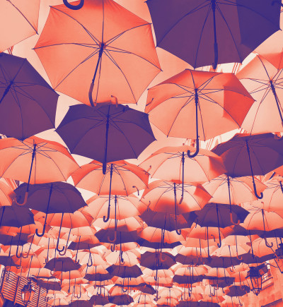 Verschiedene, geöffnete Regenschirme in warmen Farben hängen in mehreren Reihen unter einem Überhang.