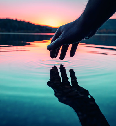 Hand über Wasser, Finger berühren die Oberfläche und erzeugen Wellen. Landschaft mit Bäumen und sanfter Himmel im Hintergrund.