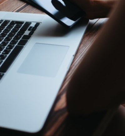 Schreibtisch mit Laptop, Person mit Handy in der Hand