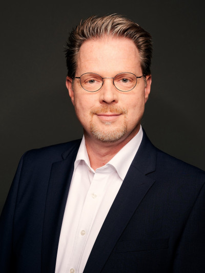 Leslie Romeo, Mann mit grauen Haaren und Brille, trägt dunklen Anzug und helles Hemd, neutraler Gesichtsausdruck.