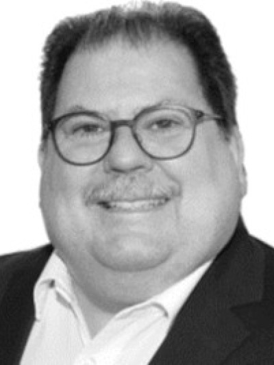 Markus Schuster, lächelnder Mann in dunkler Jacke und weißem Hemd, mit runder Brille vor hellem Hintergrund.