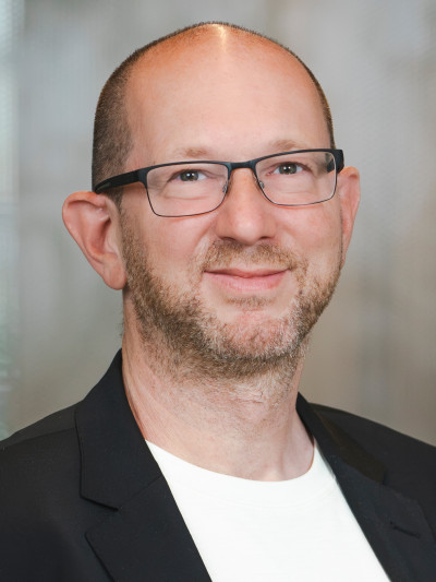 Andreas Streim, Person mit Brille und Bart, trägt schwarzen Blazer über weißem Oberteil vor unscharfem Hintergrund.