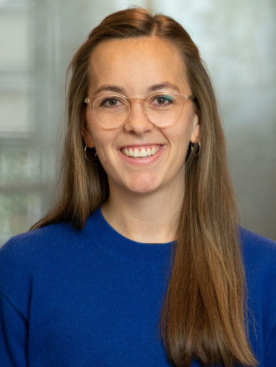 Pauline Bombeck stands in a bright room, wearing a blue sweater and glasses, smiling with an open posture.