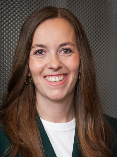 Frau mit langen, braunen Haaren lächelt in dunkelgrünem Blazer vor metallischer, strukturierter Oberfläche.