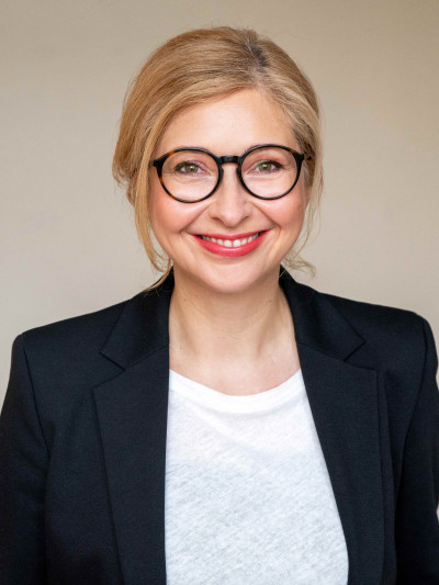 Katrin Kahle, blonde Haare im Dutt, schwarze Jacke, weißes Oberteil, Brille, lächelt vor hellem Hintergrund.