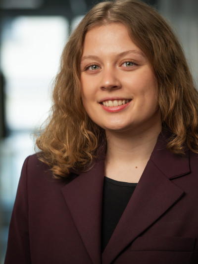 Junge Frau mit lockigem, schulterlangem Haar in dunkelviolettem Blazer, lächelt vor unscharfem, hellem Hintergrund.