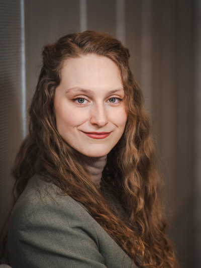 Junge Frau mit langen, lockigen Haaren in grauem Blazer, stehend an strukturierter Wand, Blick zur Kamera.
