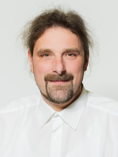 Mann mit langen, lockigen Haaren und Bart, trägt weißes Hemd, lächelt in die Kamera, hellfarbiger Hintergrund.