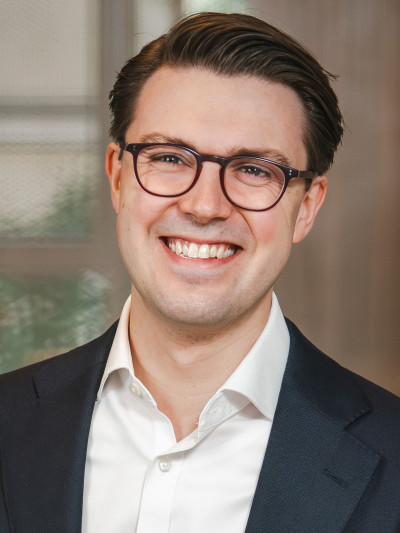 Person mit kurzen, dunklen Haaren und Brille, in hellem Hemd und dunklem Blazer, lächelt vor unscharfem Hintergrund.