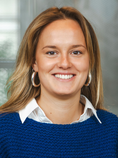 Person mit langen, hellen Haaren, blauem Pullover, weißem Hemd und runden Ohrringen, lächelt vor unscharfem Hintergrund.